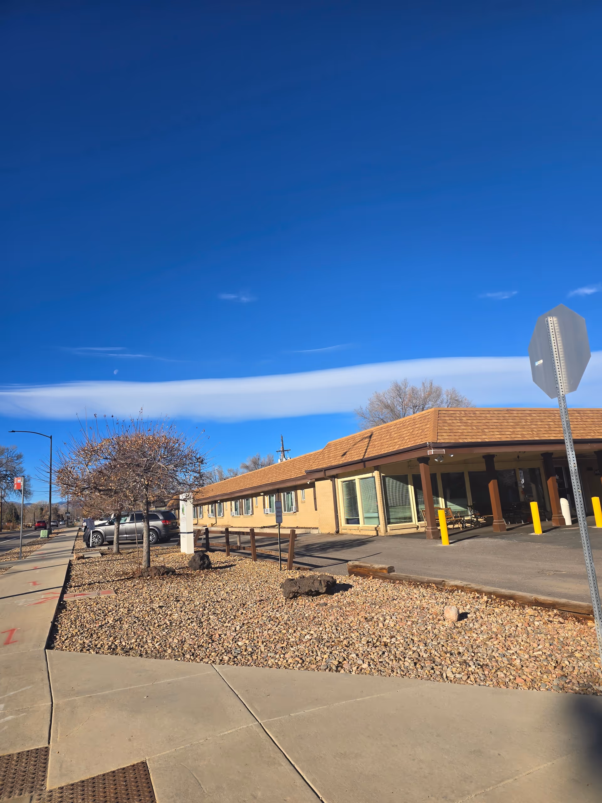 Exterior view of Mapleton Care Center building under a clear blue sky with a few clouds. The building has a brown roof and beige walls with several windows. There is a sidewalk and a landscaped area with rocks and small leafless trees in front of the building. A stop sign is visible on the right side of the image.