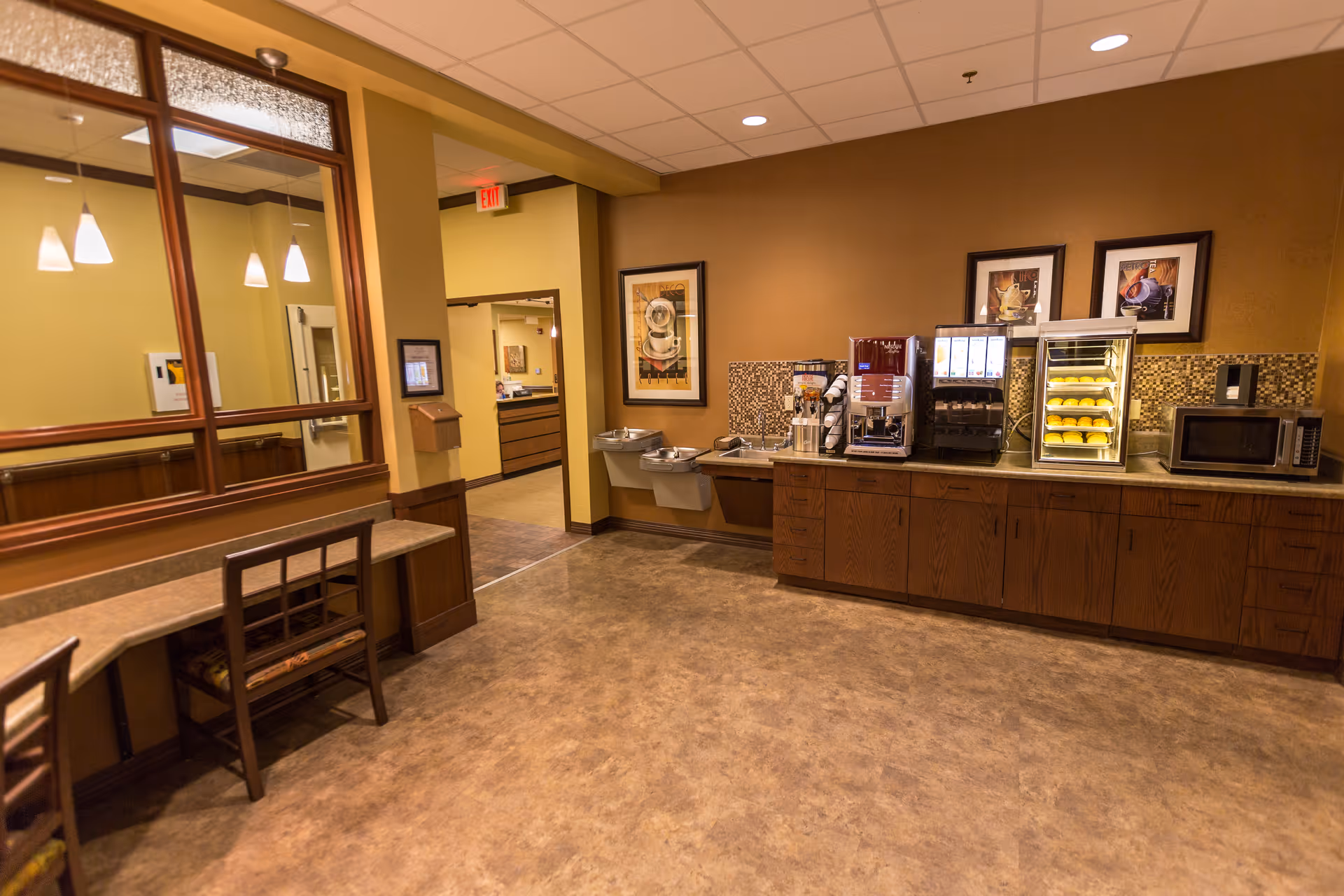 Interior view of a common area in a senior living facility featuring a countertop with a coffee machine, a microwave, a heated display case with food items, and two water fountains. There are framed pictures on the wall above the countertop and a seating area with chairs and a long desk along the opposite wall. The room has warm lighting and a tiled floor.
