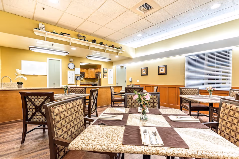 Bright dining room with set tables, patterned chairs, floral centerpieces, and a serving counter in the background.