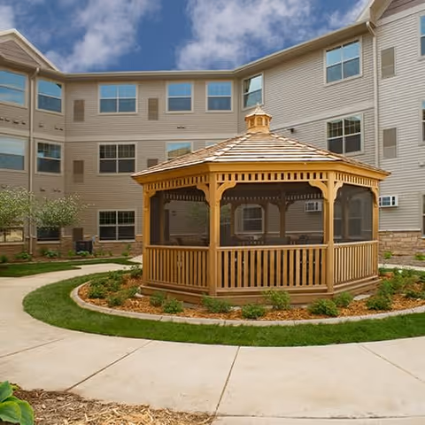 A wooden gazebo with a shingled roof is situated in the center of a circular landscaped area with grass and small plants. The gazebo is surrounded by a concrete walkway. In the background, there is a multi-story beige building with many windows under a partly cloudy sky.