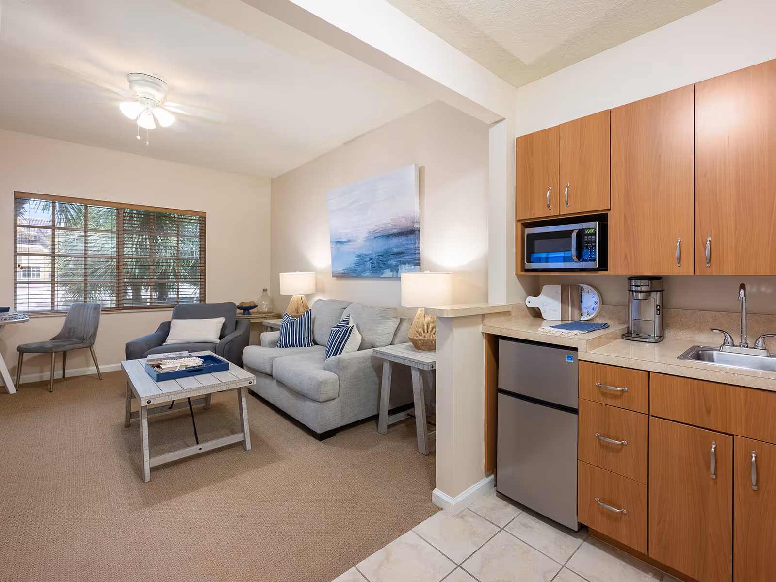 Bright open-plan living area with a sofa, armchair, coffee table and wall art, adjacent to a small kitchenette with wooden cabinets and a mini-fridge.
