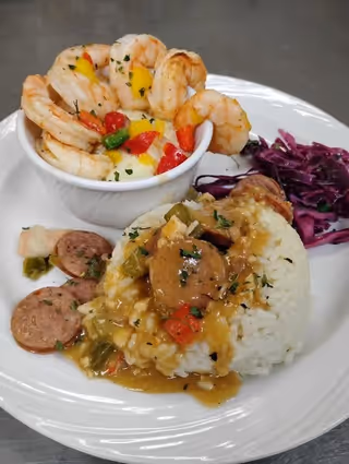 A plate of food featuring a mound of white rice topped with sausage and a savory sauce with diced vegetables, a side of cooked purple cabbage, and a small bowl filled with shrimp and diced colorful vegetables.