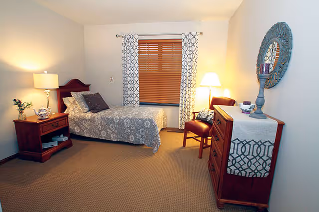A cozy bedroom with a single bed covered in a floral patterned bedspread, a wooden nightstand with a lamp and decorative items, a window with wooden blinds and patterned curtains, a wooden dresser with a decorative runner and a round mirror above it, and a chair with a cushion next to a floor lamp.