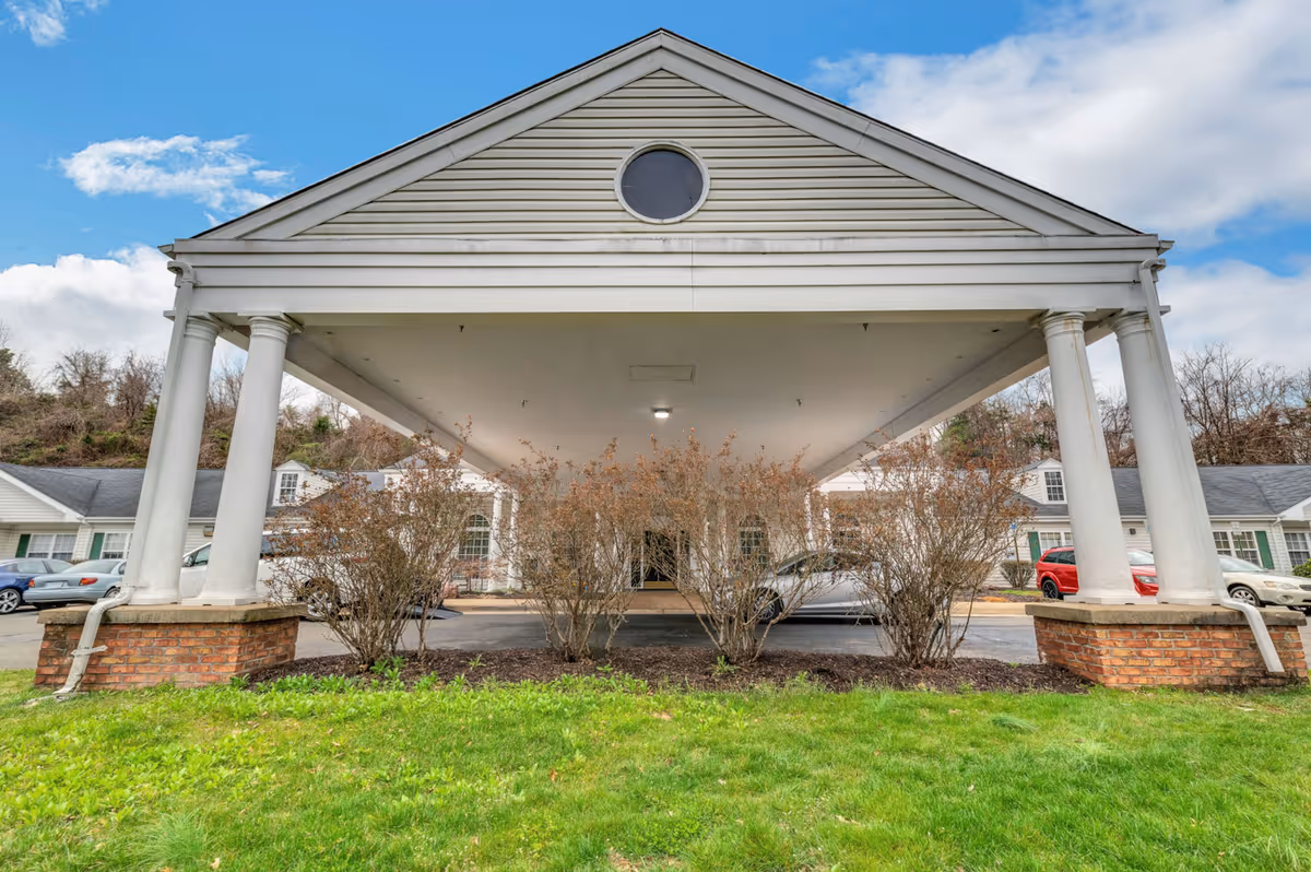 Front exterior view of a senior living facility entrance with a covered driveway supported by white columns and brick bases, surrounded by bushes and green grass under a partly cloudy sky.