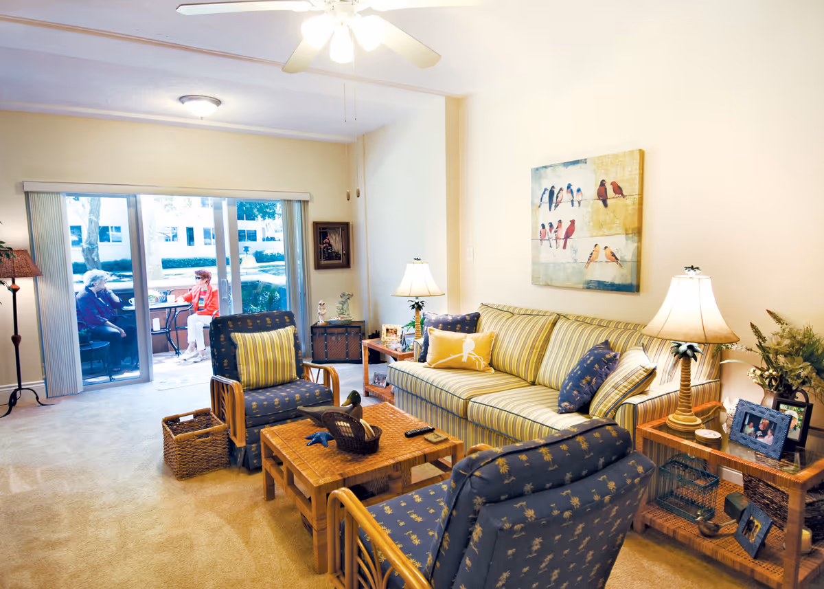 Bright living room with a striped sofa, patterned armchairs, coffee table and lamps, with sliding glass doors showing two people seated outside.