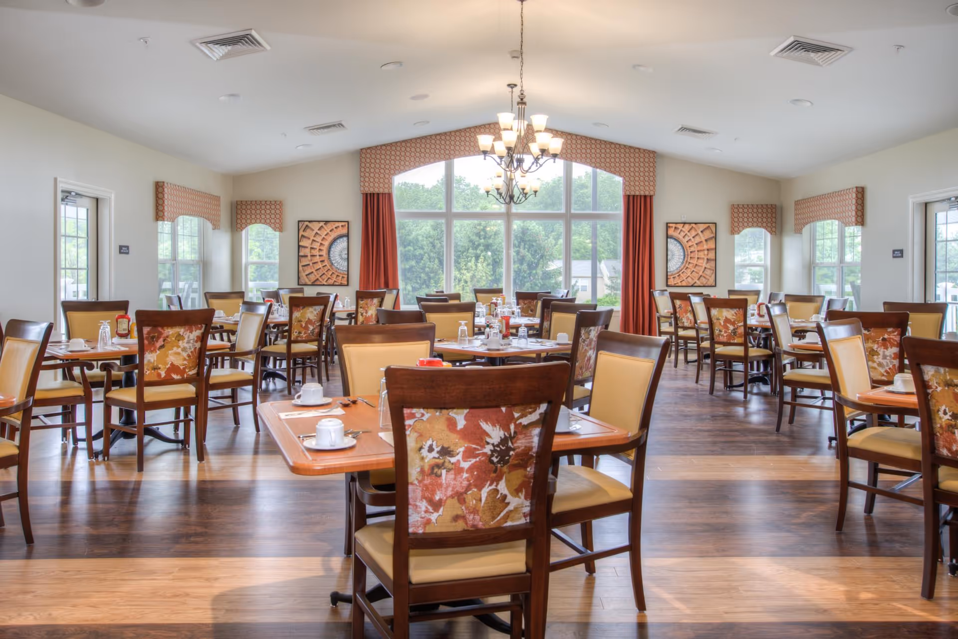 A bright dining room with multiple wooden tables and chairs featuring floral upholstery. The room has large windows with red curtains and valances, allowing natural light to fill the space. Tables are set with cups, saucers, and condiments. The floor is wooden, and there are decorative wall hangings on the far wall.