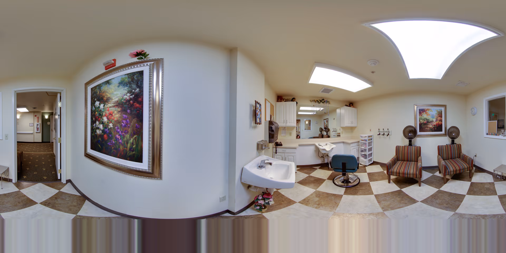Interior view of a senior living facility's hair salon area with checkered brown and beige floor tiles, a white sink, a salon chair in front of a mirror, white cabinets, two striped armchairs with hair dryers behind them, and colorful floral paintings on the walls.