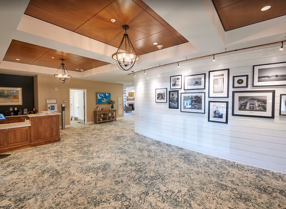 Interior view of a senior living facility lobby or common area with a patterned carpet, wooden reception desk on the left, a wall decorated with framed black and white photographs on the right, and two hanging light fixtures on the ceiling.