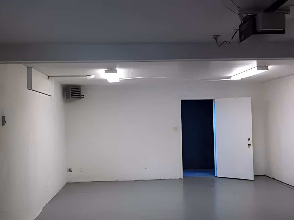 Empty room with white walls and ceiling, gray floor, fluorescent ceiling lights, a wall-mounted heater or vent, and a partially open white door revealing a dark blue room beyond.