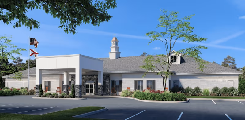 Exterior view of a single-story senior living facility building with a covered entrance, stone and brick facade, landscaped bushes and trees, and an empty parking lot under a clear blue sky.