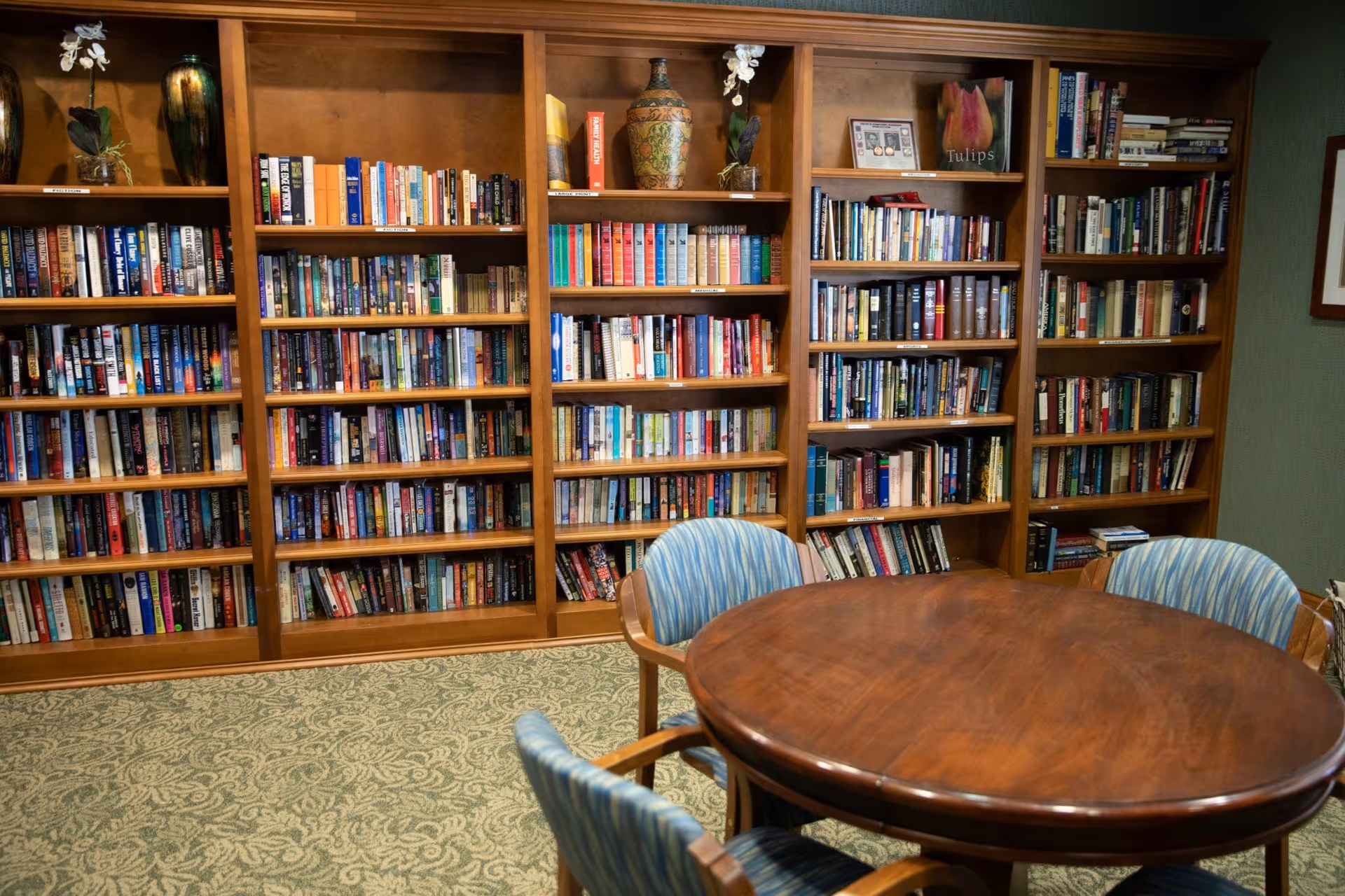Wooden bookshelves filled with books line the wall behind a round wooden table and upholstered chairs in a reading room.