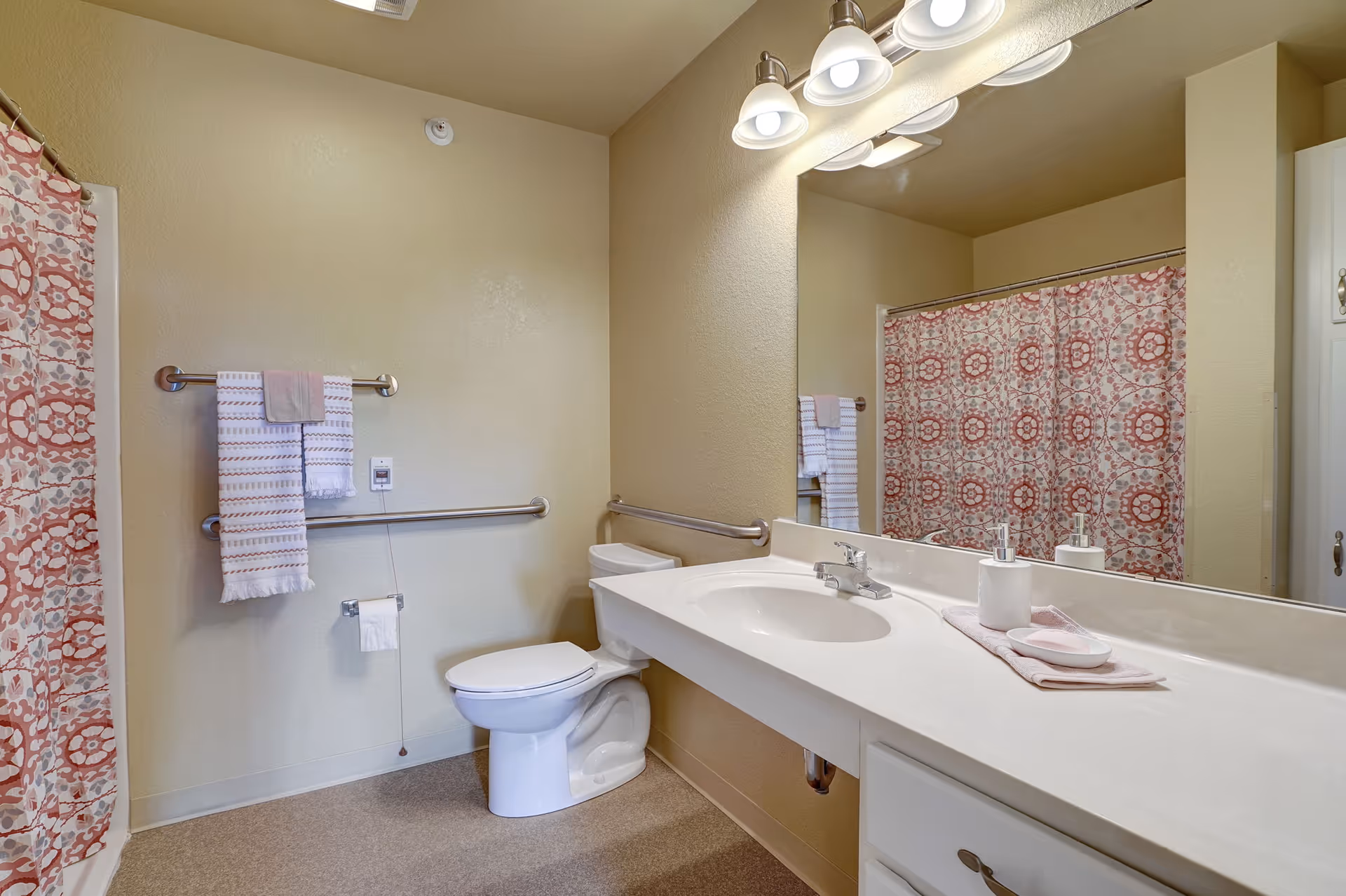 A clean and accessible bathroom with a white toilet, a long white countertop with a sink, a large mirror above the sink, and a shower with a patterned red and white curtain. There are grab bars near the toilet and a towel rack with folded towels on the wall.