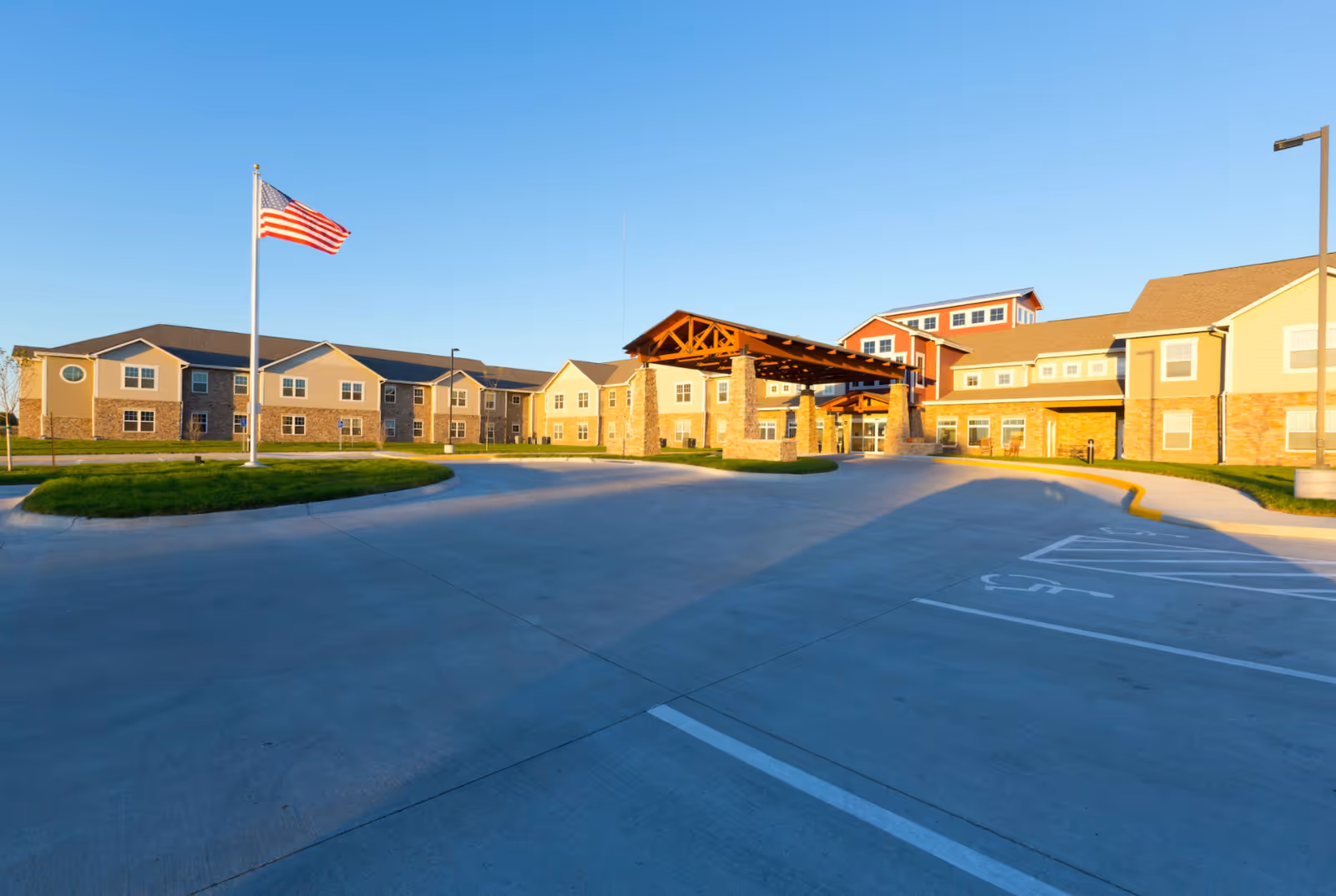 Front entrance of a multi‑story senior living building with a covered porte‑cochere, circular driveway, and an American flag on a flagpole under a clear blue sky.