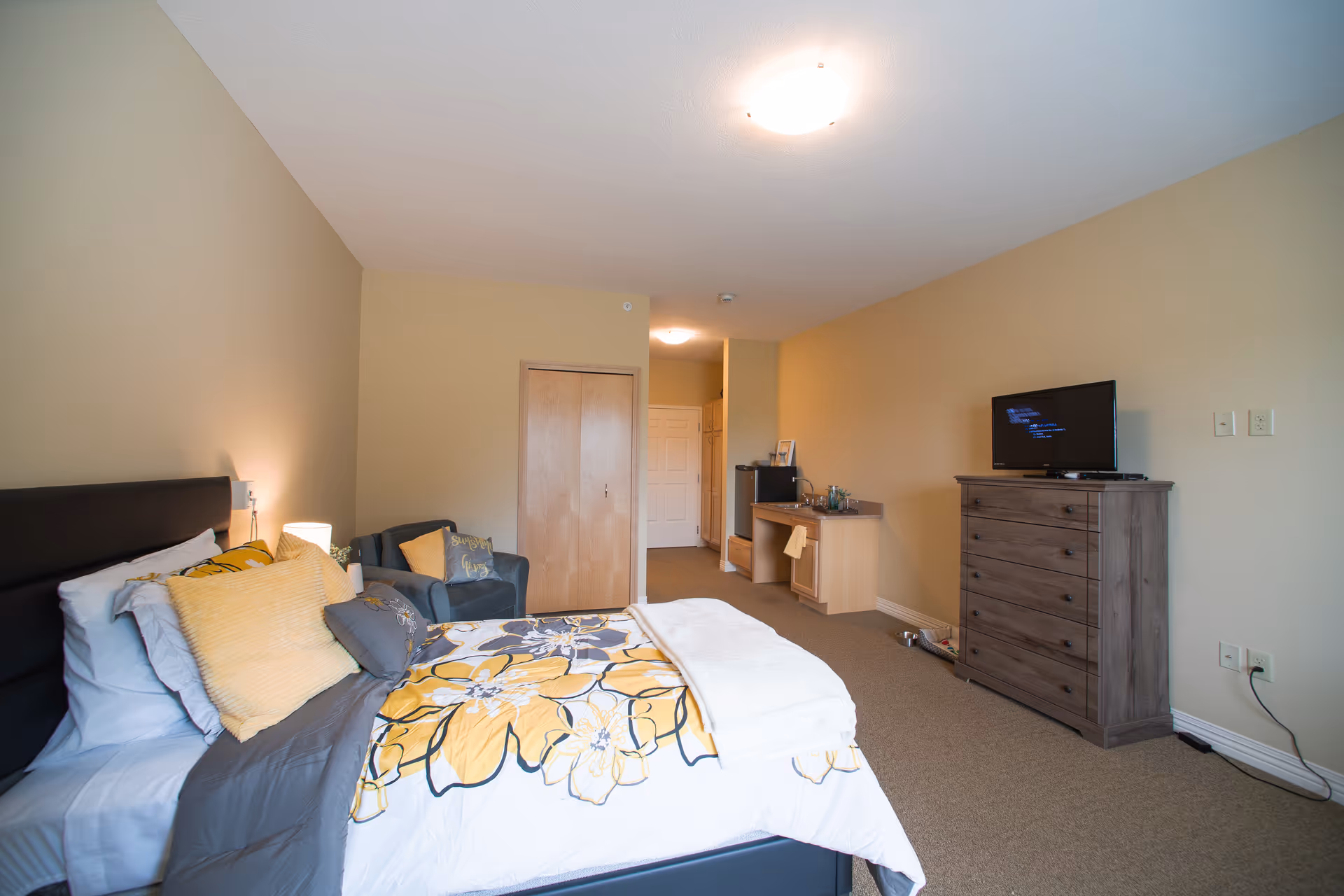 A senior living facility bedroom with a bed featuring yellow and gray floral bedding, a gray armchair with yellow pillows, a wooden dresser with a TV on top, and a small kitchenette area with a sink and mini fridge. The walls are beige and the carpet is light brown.