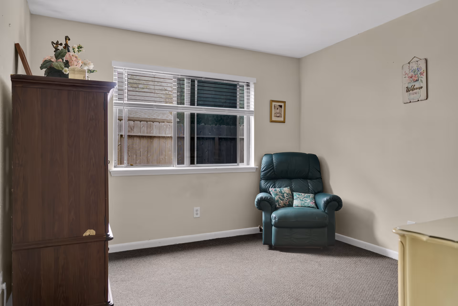 A small room with beige walls and carpeted floor featuring a green leather recliner chair with two floral cushions, a wooden cabinet with decorative flowers on top, a window with white blinds showing a wooden fence outside, and two small framed wall decorations.