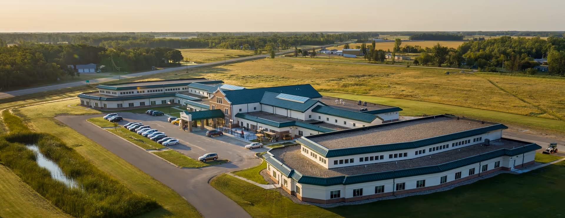 Aerial view of a multi-wing senior living campus with green roofs, a parking lot, driveways and surrounding open fields.