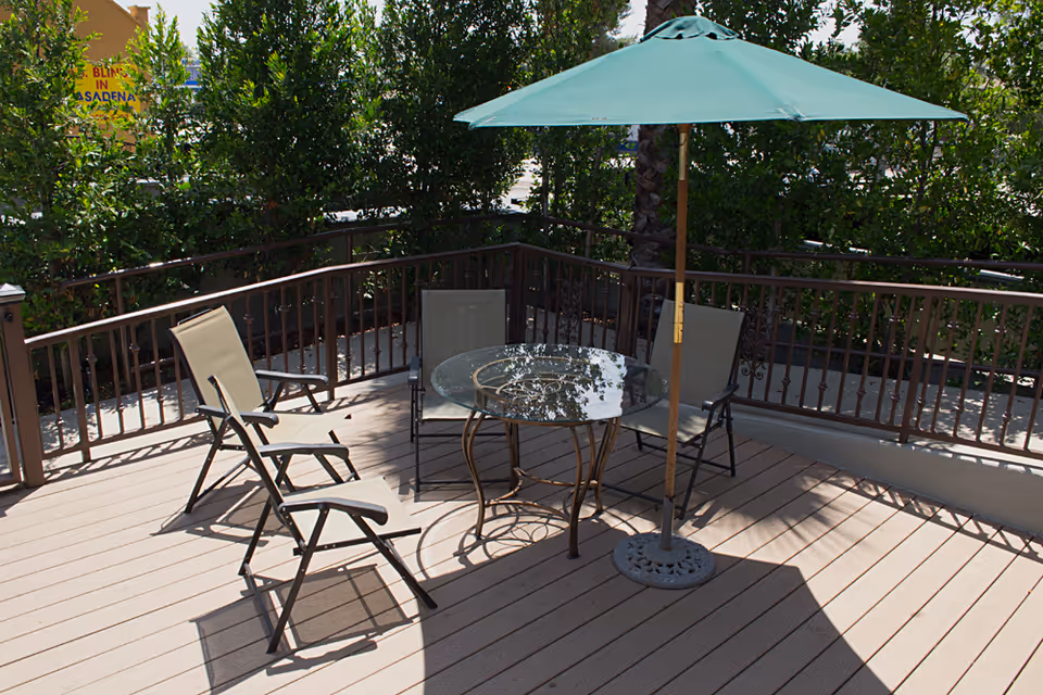 Outdoor patio area with a round glass table, four beige chairs, and a green umbrella providing shade. The patio is surrounded by a brown railing and lush green bushes.