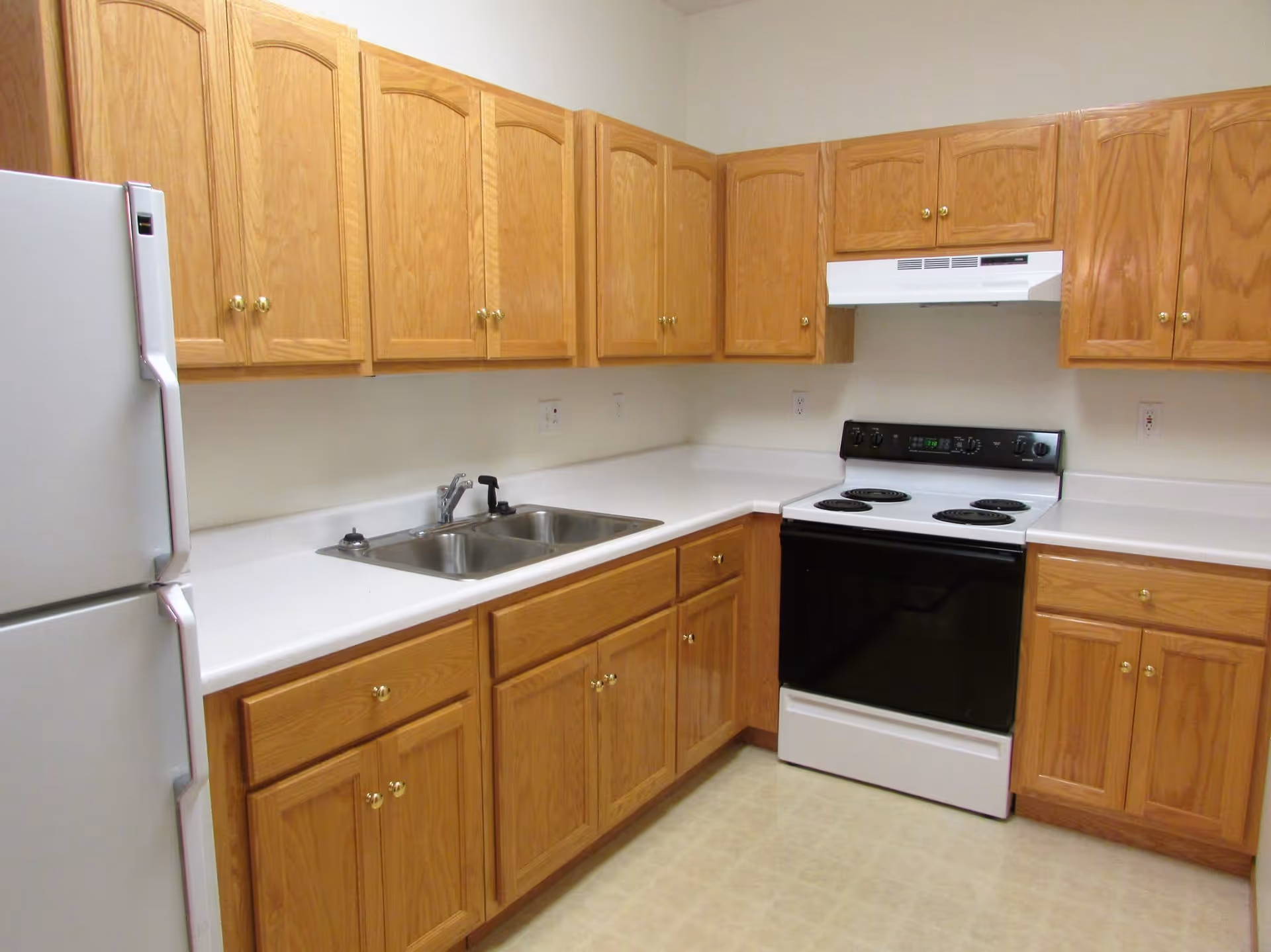 A kitchen with wooden cabinets, a white refrigerator, a double stainless steel sink, a white electric stove with a black oven door, and a white range hood. The countertops are white, and the floor has a light-colored tile pattern.