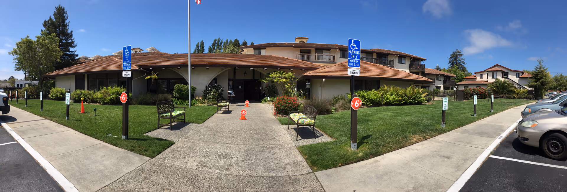 Panoramic view of the front exterior of La Posada Retirement Community (Independent Living) building with a tiled roof, green lawn, benches, handicap parking signs, and parked cars under a clear blue sky.