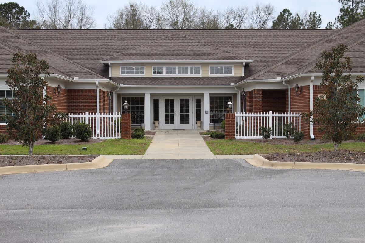 Front exterior view of The Summit of Atmore assisted living facility showing a single-story brick building with a peaked roof, white trim, and a central entrance with double glass doors. The entrance is flanked by white picket fences and small landscaped areas with shrubs and trees.