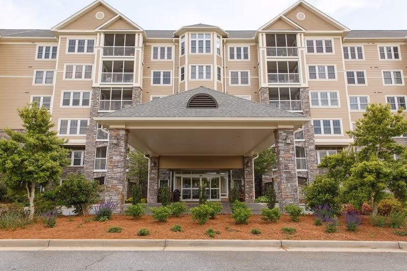 Front exterior of a multi-story senior living building with a covered drop-off entrance and landscaped beds.