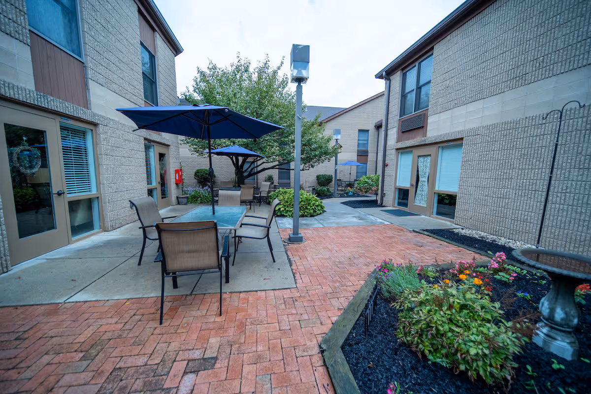 Outdoor courtyard area at Independence Court of Quakertown with brick and concrete pathways, patio tables with umbrellas, chairs, landscaped flower beds, and surrounding two-story building walls with windows and doors.