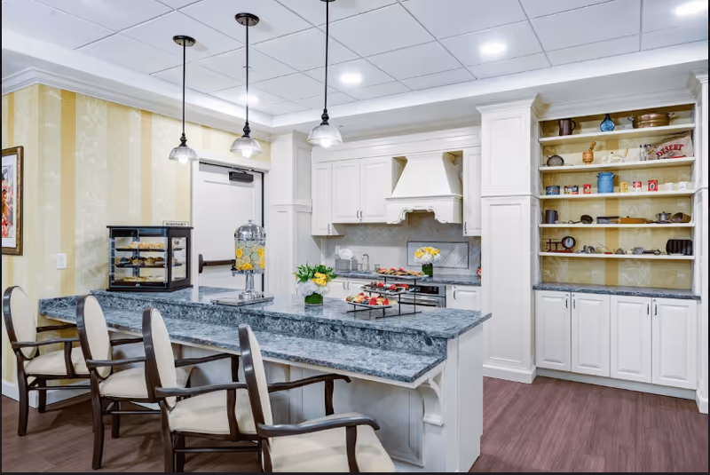 A bright and clean kitchen area with a long marble countertop island surrounded by four cushioned chairs. The kitchen features white cabinetry, a stove, and a range hood. On the countertop, there are trays with desserts, a glass beverage dispenser with lemon water, and small flower arrangements. To the right, there is a built-in shelving unit with various decorative items and kitchenware. The room has pendant lights hanging from the ceiling and wood flooring.