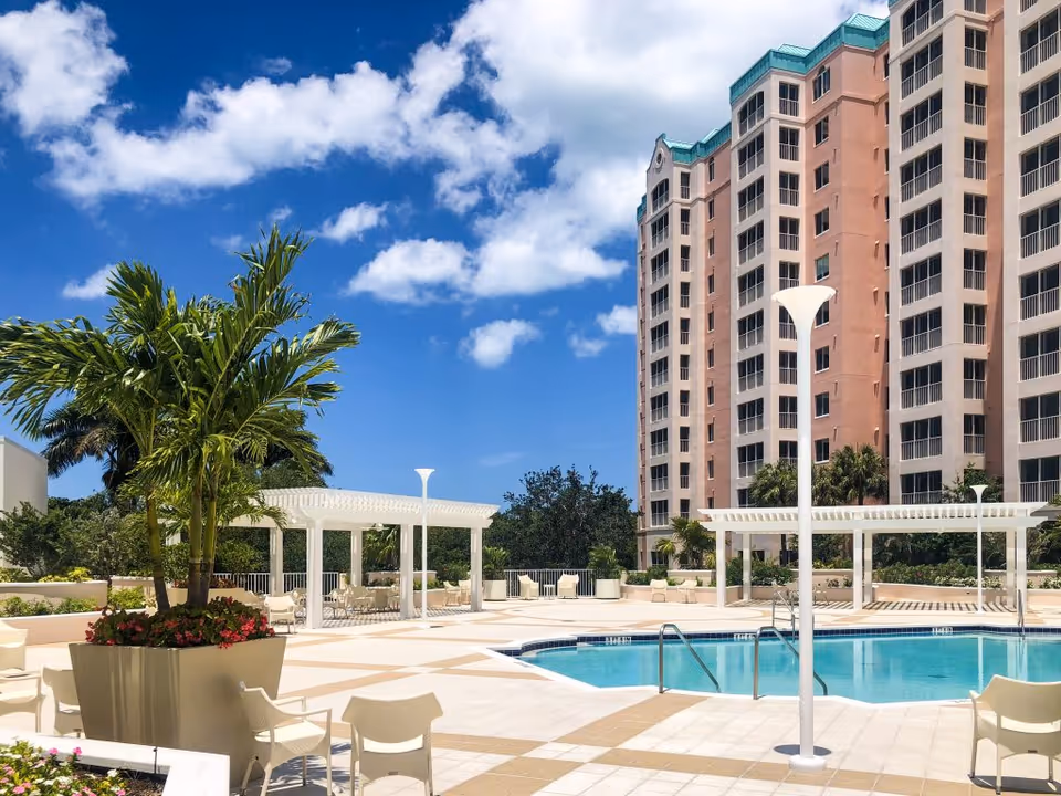 Sunlit outdoor pool deck with lounge chairs, pergolas, palm plants, and a tall residential building under a blue sky.
