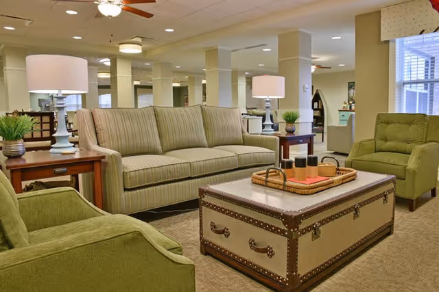 A cozy living room area in a senior living facility featuring a striped beige sofa, two green armchairs, a large trunk-style coffee table with a tray holding decorative items, two side tables with lamps, and a ceiling fan above. The room has beige walls and carpet, with large windows allowing natural light.