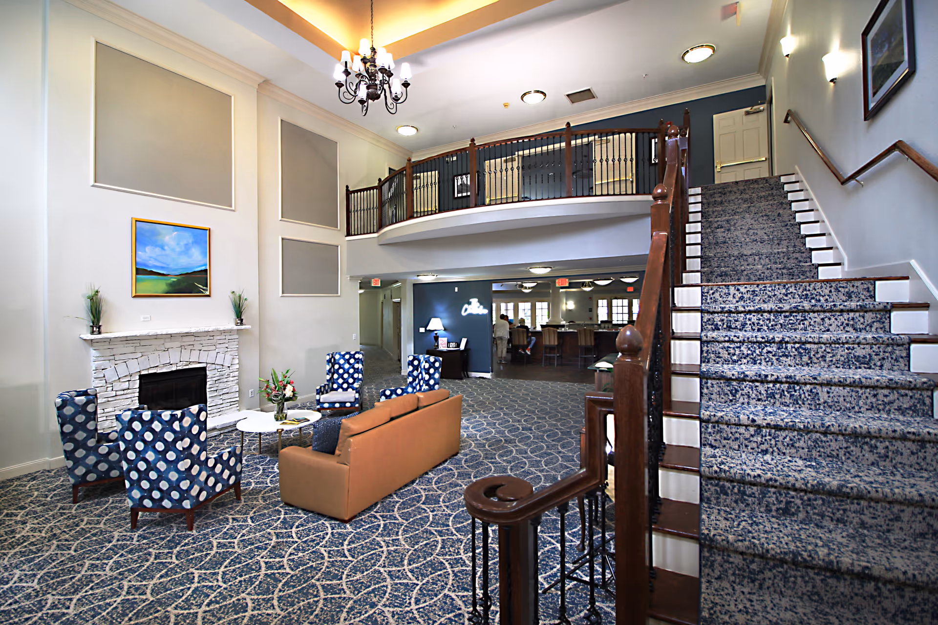 Interior view of a senior living facility lounge area with a staircase on the right, patterned carpet, a stone fireplace with a landscape painting above it, a chandelier hanging from the ceiling, and seating including a brown sofa and blue patterned armchairs arranged around a white coffee table.