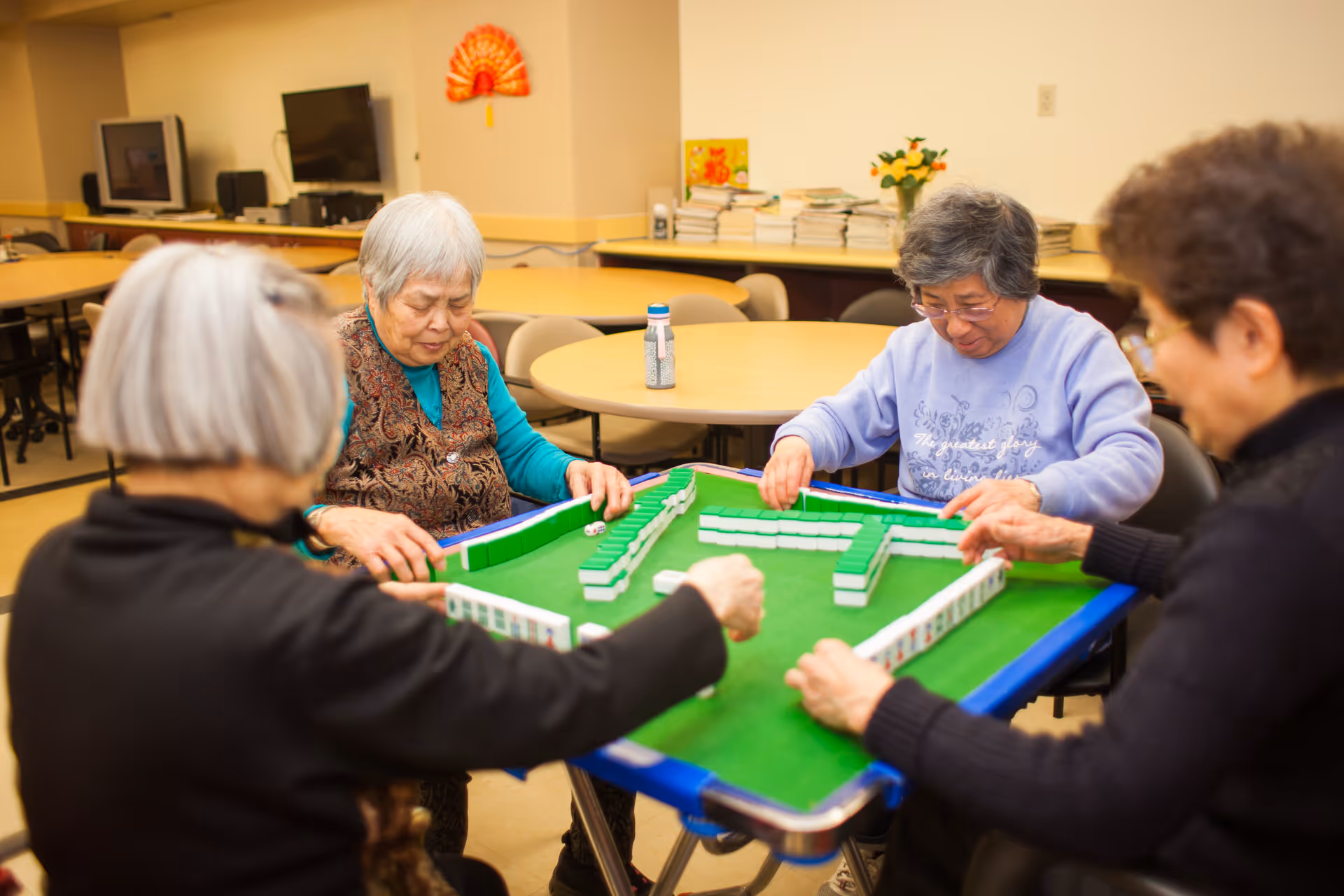Four elderly women sitting around a table playing a game of Mahjong in a well-lit room with round tables and chairs in the background.
