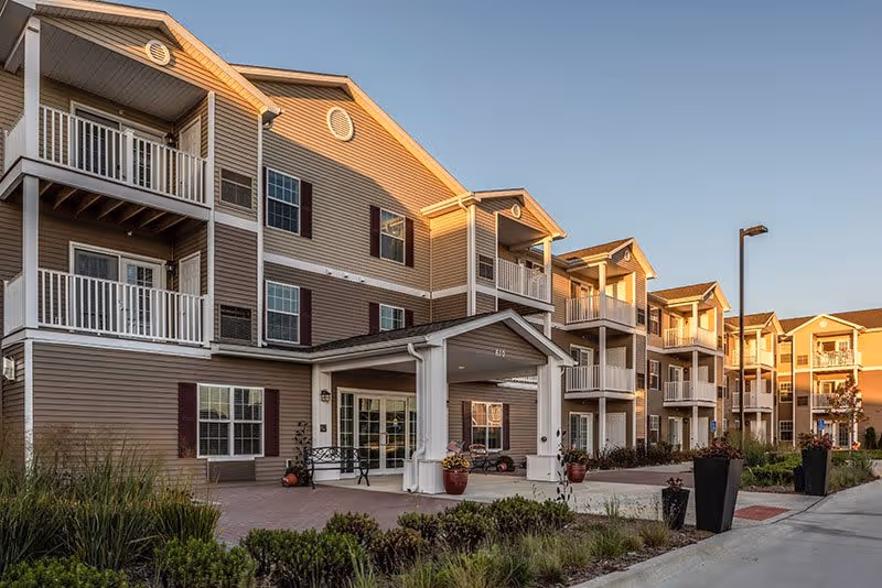 Exterior view of a multi-story senior living facility building with balconies, a covered entrance, and landscaped greenery under a clear sky during sunset.