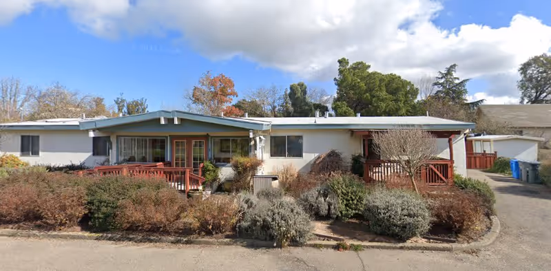 Single-story building with a light-colored exterior and a blue roof, featuring two wooden porch areas with railings. The front yard has various bushes and shrubs, and there are trees in the background under a partly cloudy sky.