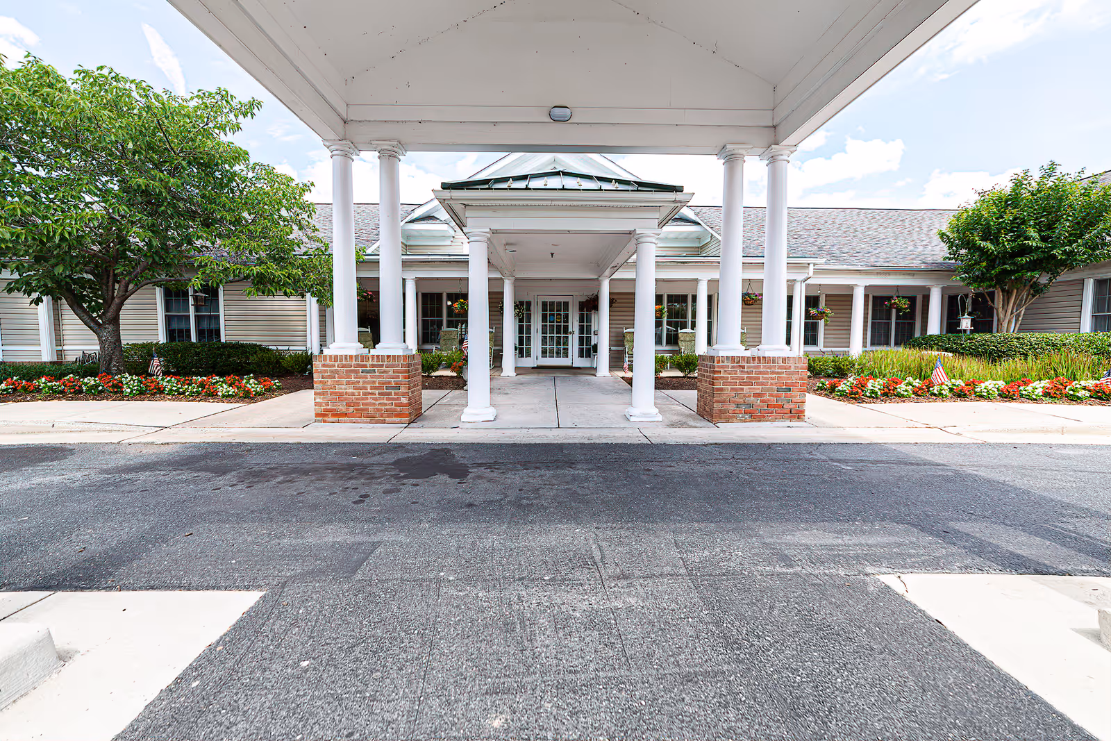Front entrance of HeartFields Assisted Living at Easton showing a covered driveway with white columns and brick bases, surrounded by well-maintained landscaping including trees and flower beds.