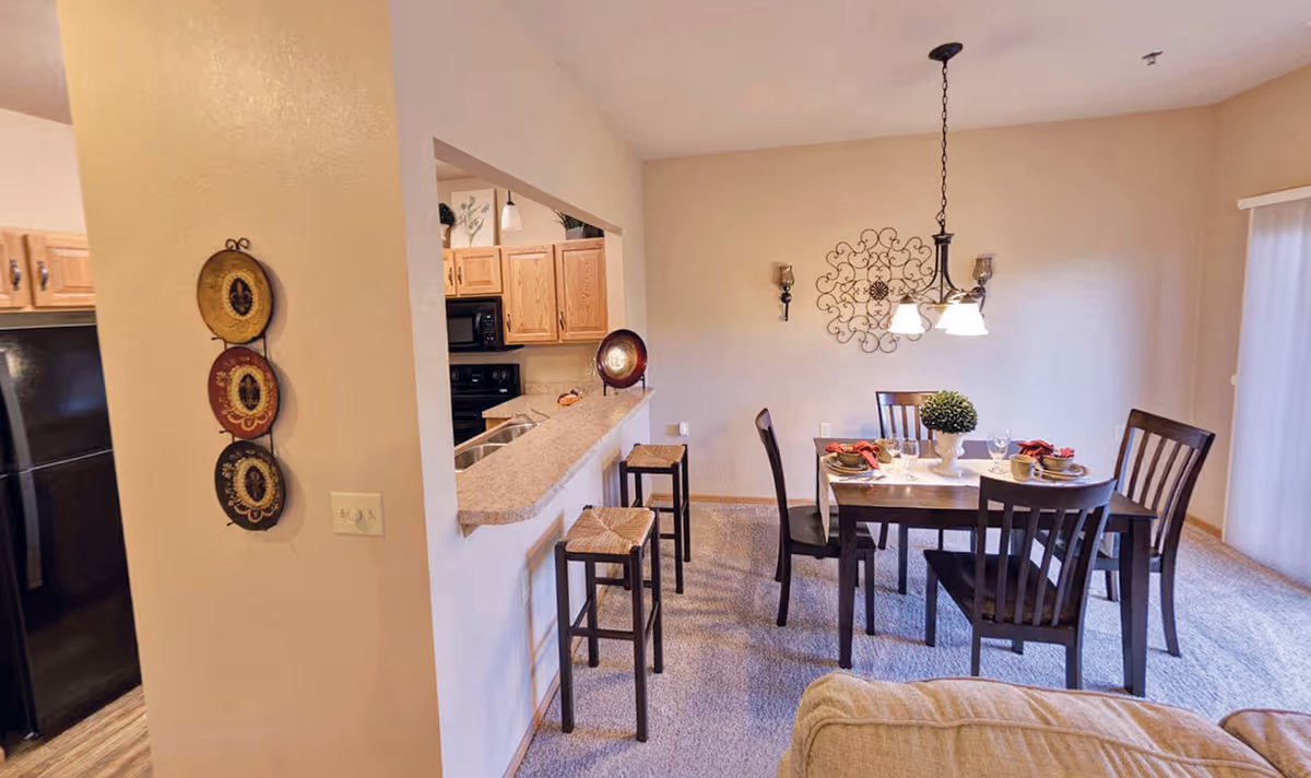Interior view of a senior living facility showing a dining area with a dark wooden table set for four, surrounded by matching chairs. Adjacent to the dining area is a kitchen with wooden cabinets, a black refrigerator, and a countertop with two bar stools. The walls are light-colored, and there is decorative wall art and a hanging light fixture above the dining table.