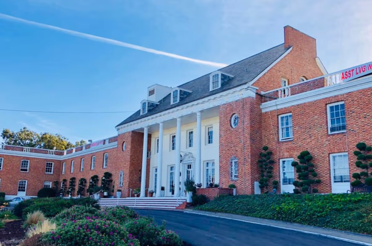 Exterior view of a large brick building with white columns at the entrance, manicured bushes, and a clear blue sky with a visible contrail.