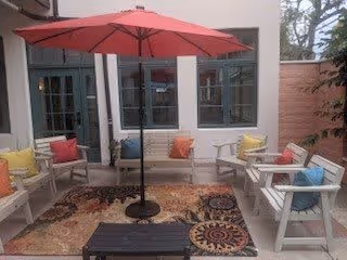 Outdoor patio area with white wooden chairs and benches arranged around a large red umbrella. The seating has colorful cushions in yellow, red, blue, and orange. A patterned rug is placed under the seating area, and there is a small black table in the center. The patio is adjacent to a building with large windows and a wooden privacy fence on one side.