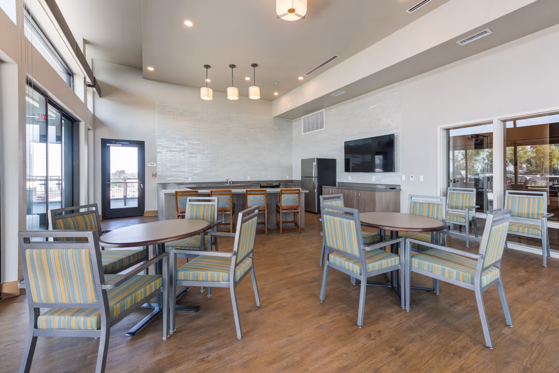 Bright communal dining/lounge area with round tables and striped chairs, a kitchenette and wall-mounted TV.