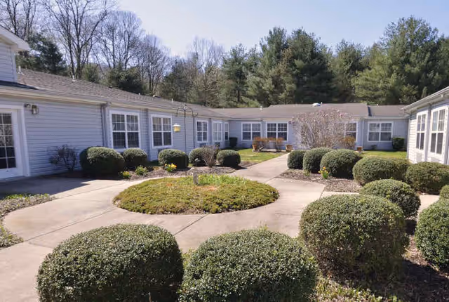 Sunlit central courtyard of a single-story building with a circular walkway, trimmed shrubs, benches and surrounding windows.