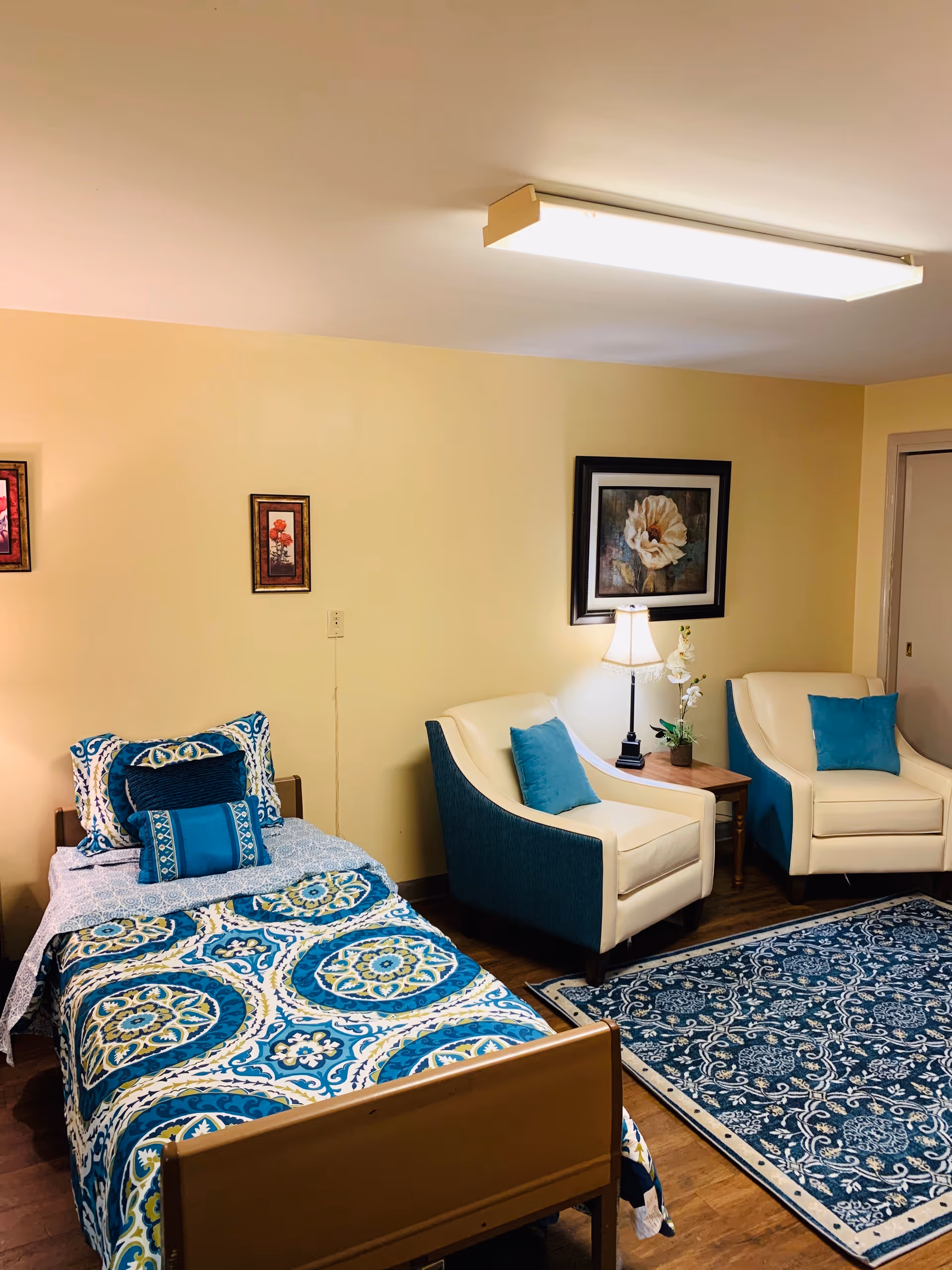 A cozy room with a single bed covered in a blue, white, and yellow patterned bedspread and matching pillows. Next to the bed are two cream and blue armchairs with blue cushions, a wooden side table with a lamp and a small plant. The walls are painted light yellow and decorated with framed floral artwork. A blue patterned rug covers part of the wooden floor.