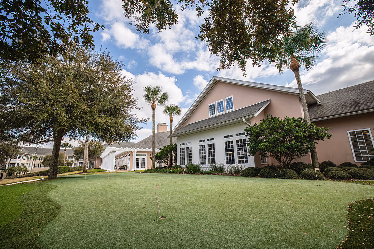 Outdoor view of Marsh's Edge facility showing a putting green with several holes, surrounded by trees and shrubs. The building has large windows and a sloped roof under a partly cloudy sky.
