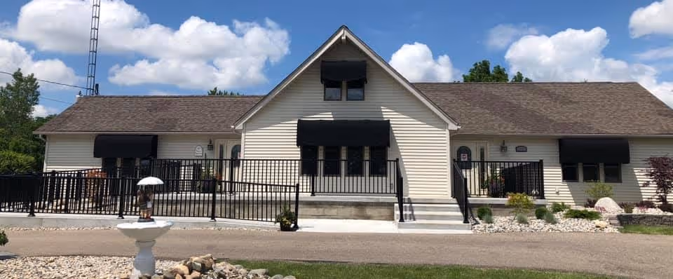 Exterior view of a single-story building with beige siding and a brown shingled roof under a blue sky with clouds. The building has black awnings over the windows and doors, a black metal railing along the front steps and ramp, and a small landscaped area with rocks and plants in front.