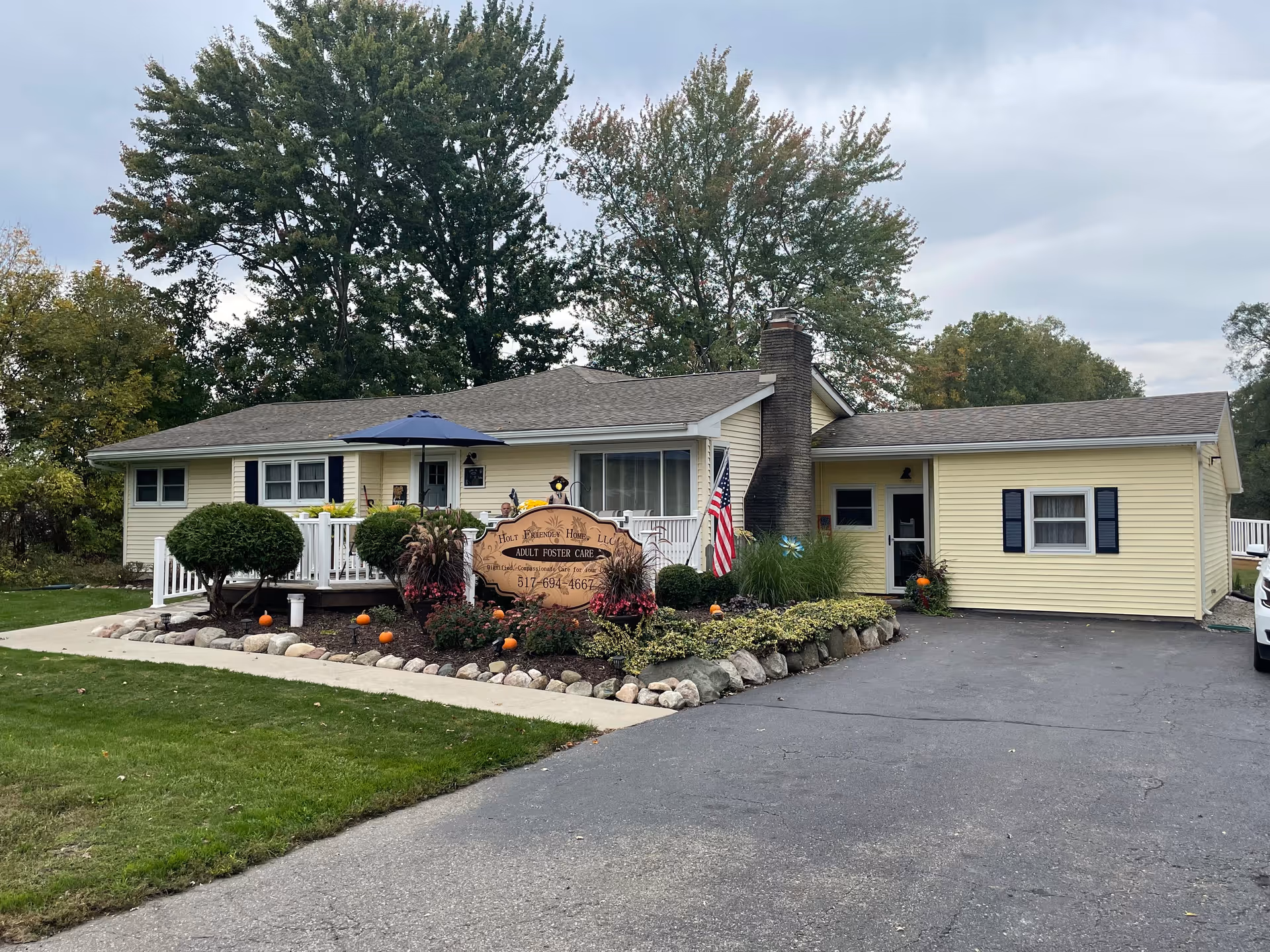 Single-story yellow building with a gray roof, surrounded by landscaped bushes and plants. A wooden sign in front reads 'Holt Friendly Home, LLC Adult Foster Care' with a phone number. An American flag is displayed near the entrance, and there is a paved driveway leading up to the building. Trees are visible in the background under a cloudy sky.