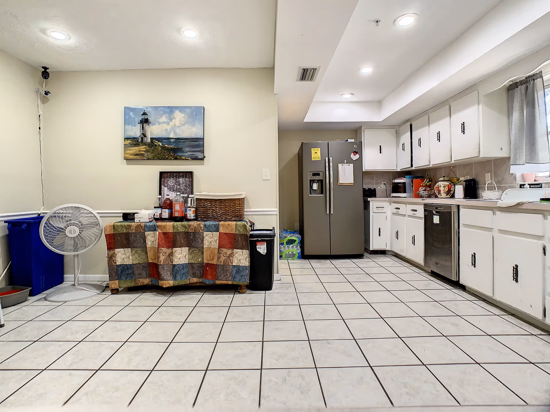 A clean kitchen area with white cabinets, a stainless steel refrigerator, and a dishwasher. There is a table covered with a colorful patchwork cloth holding various bottles and a basket. A standing fan and recycling bin are visible on the left side. A painting of a lighthouse hangs on the wall above the table. The floor is tiled with white tiles and dark grout lines.