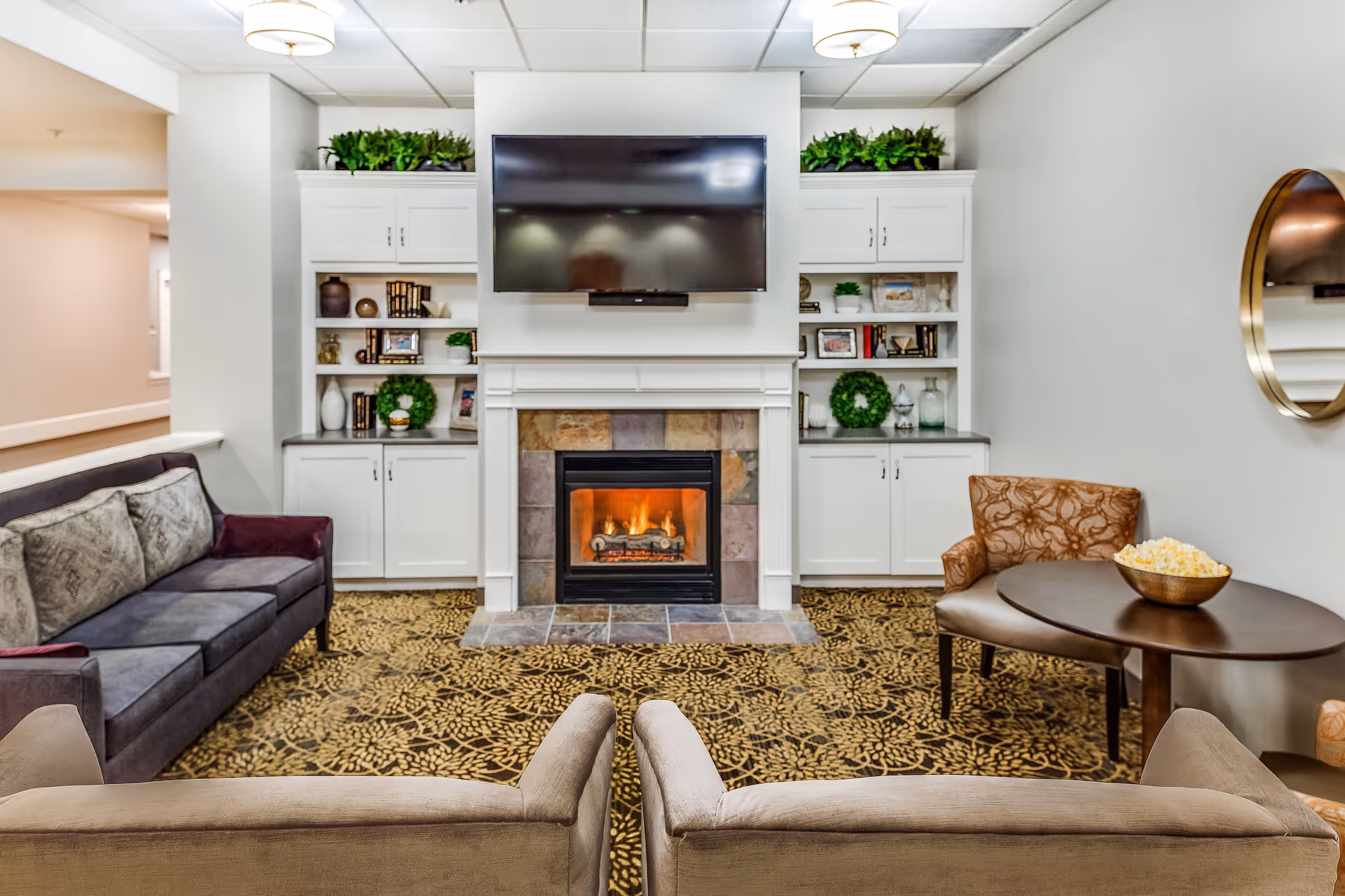 A cozy living room area with a fireplace in the center, above which is a mounted flat-screen TV. On either side of the fireplace are white built-in shelves and cabinets decorated with plants, books, and decorative items. The room features a patterned carpet, a dark gray sofa with cushions on the left, two beige armchairs facing the fireplace, and a small round table with a bowl of popcorn and an upholstered chair on the right. A round mirror hangs on the right wall.