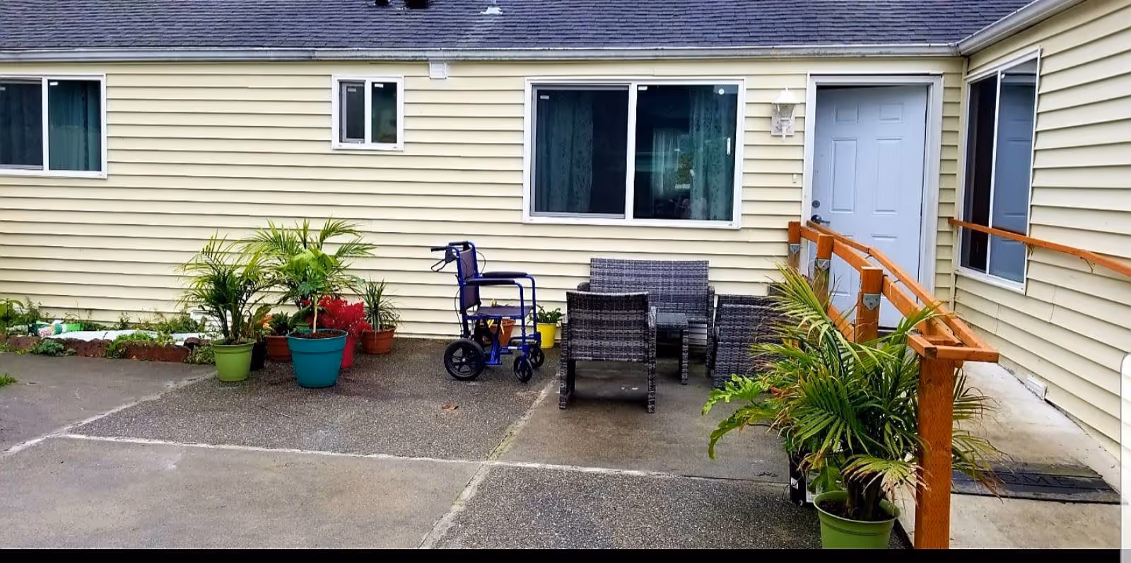 Outdoor patio area with several potted plants, a blue wheelchair, and a set of wicker chairs and a small table. The patio is adjacent to a beige building with multiple windows and a white door with a wooden handrail ramp leading to it.