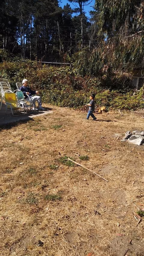 An elderly person sitting on a lawn chair on a concrete patio area next to a dry grassy yard. A young child is walking on the grass holding sticks. There are bushes and tall trees in the background under a clear blue sky.