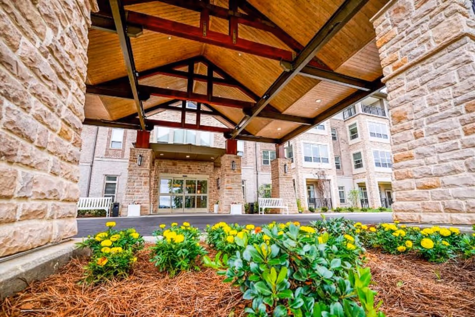 Entrance of a senior living facility with a covered driveway supported by stone pillars and wooden beams. There are yellow flowers and green shrubs planted in the foreground, and the building has multiple windows and balconies.
