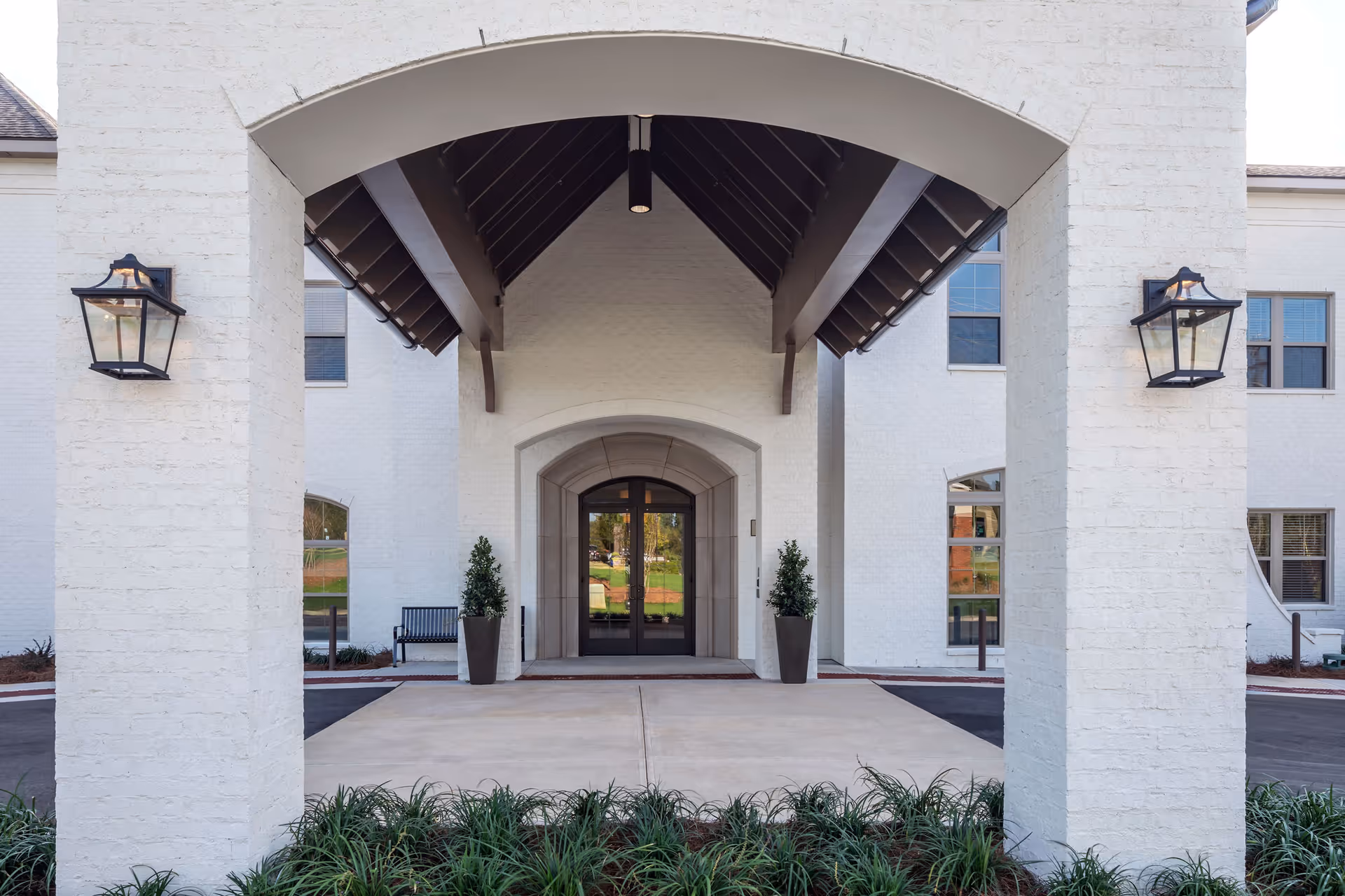 Entrance of a senior living facility with white brick walls, a covered archway supported by two pillars, two wall-mounted lantern lights, potted plants on either side of the glass double doors, and a bench visible to the left side.