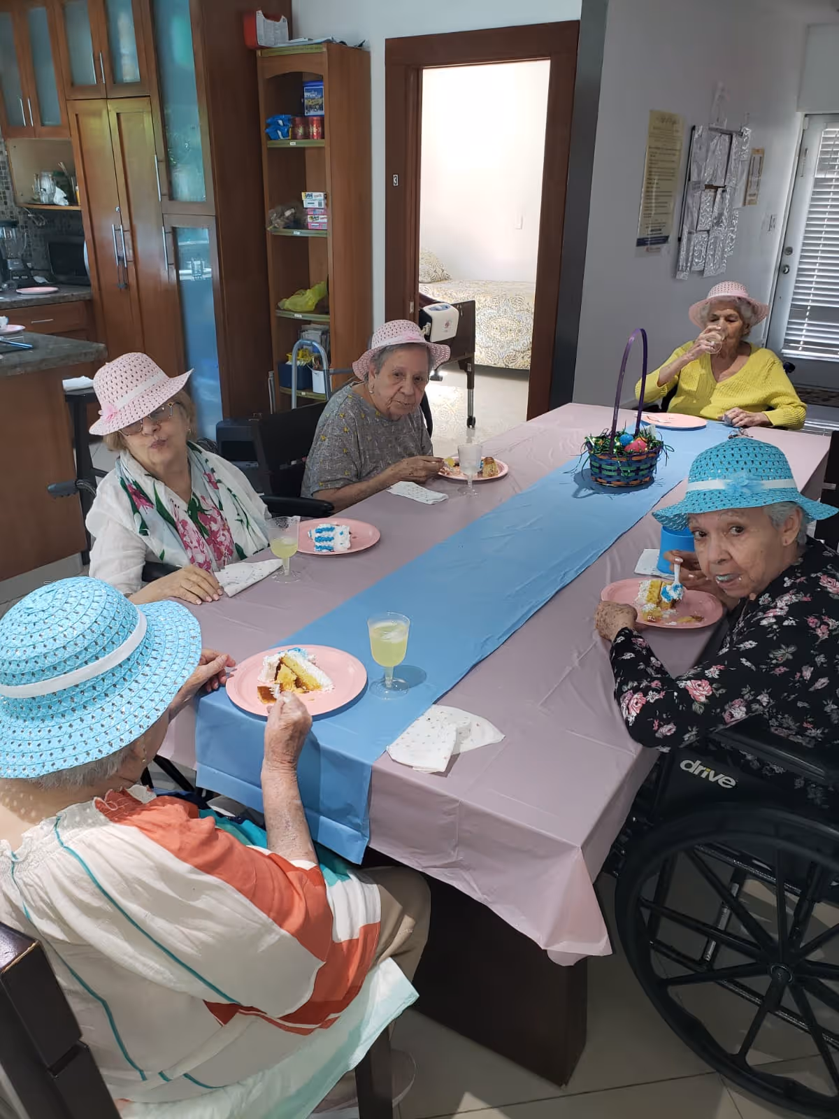 Five elderly women wearing colorful hats are seated around a dining table covered with a pink tablecloth and a blue table runner. They are eating cake and drinking a light-colored beverage. The setting appears to be a dining area in a senior living facility, with a kitchen visible in the background and a bedroom visible through an open door.
